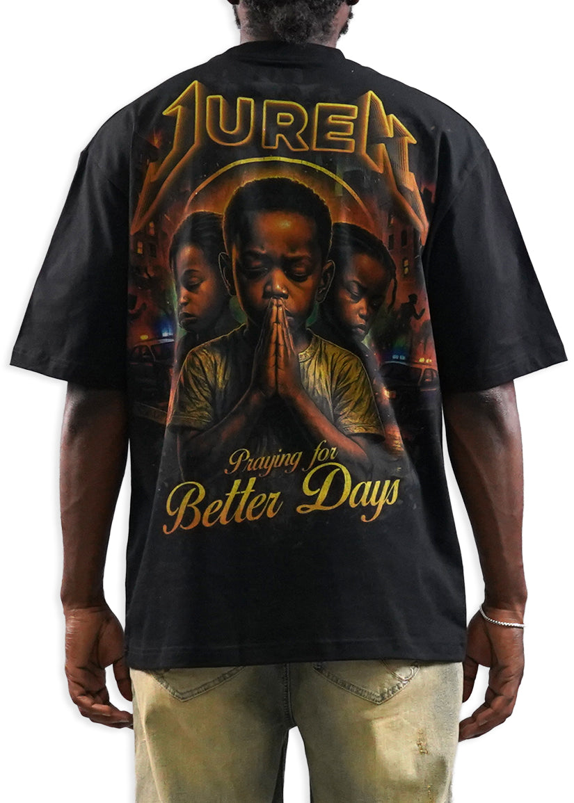 A man shown from the back wearing a black t-shirt with a large graphic print. The graphic features the word 'JUREN' in a gold metallic font at the top, a central portrait of a man in prayer, and the phrase 'Praying for Better Days' in gold at the bottom.