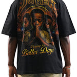 A man shown from the back wearing a black t-shirt with a large graphic print. The graphic features the word 'JUREN' in a gold metallic font at the top, a central portrait of a man in prayer, and the phrase 'Praying for Better Days' in gold at the bottom.