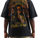 A man shown from the back wearing a black t-shirt with a large graphic print. The graphic features the word 'JUREN' in a gold metallic font at the top, a central portrait of a man in prayer, and the phrase 'Praying for Better Days' in gold at the bottom.