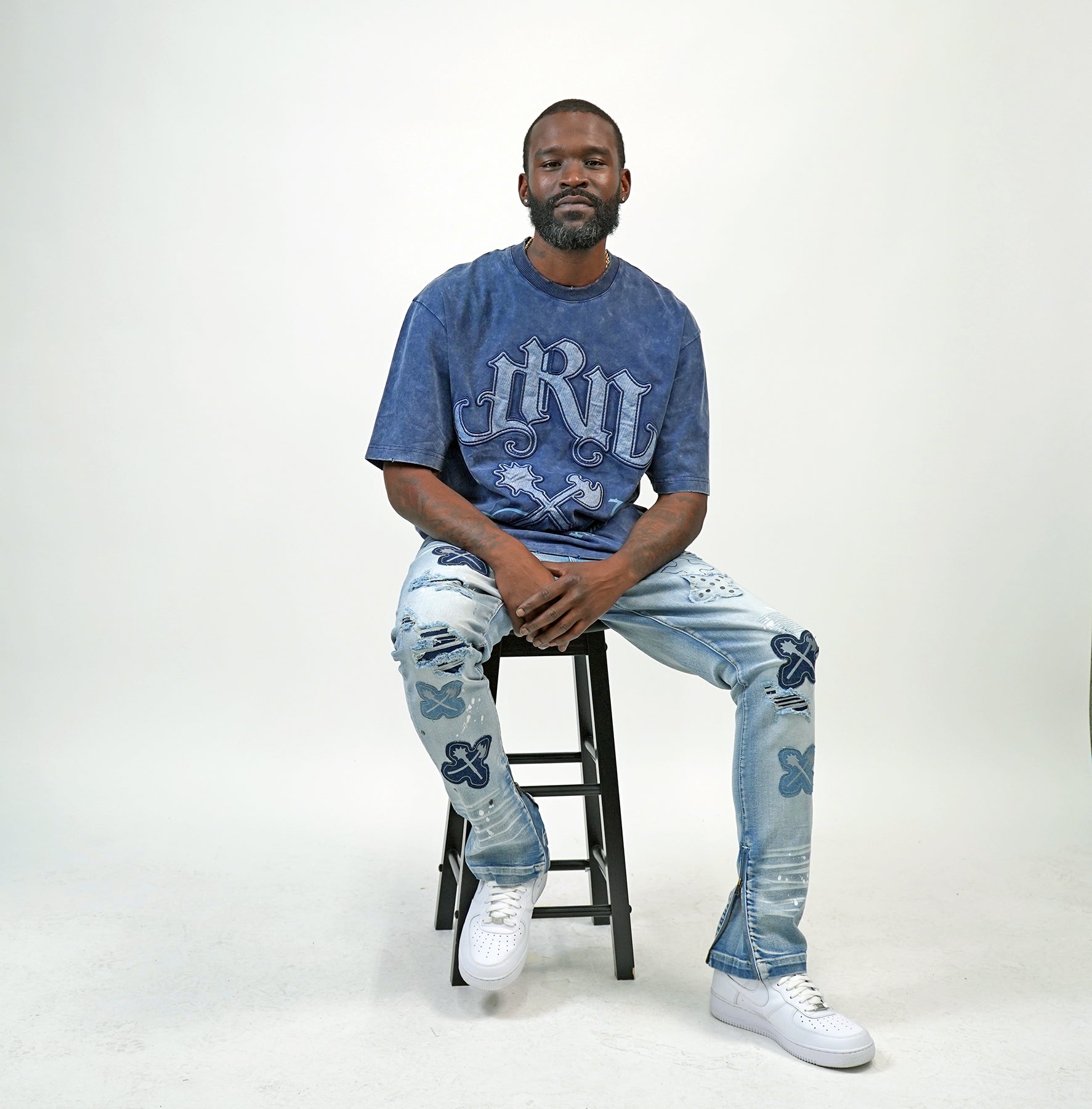 Man sitting on a stool wearing a blue graphic t-shirt and jeans, Man wearing a blue acid wash t-shirt with text and graphics on the Juren logo battle axe.