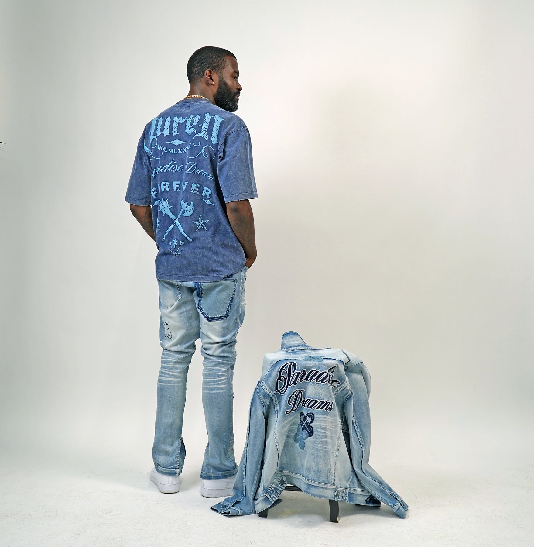 Man wearing a blue acid wash graphic t-shirt and jeans standing next to an open denim jacket on a white background