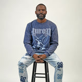 Man wearing a blue graphic sweatshirt and light blue jeans sitting on a black stool against a white background