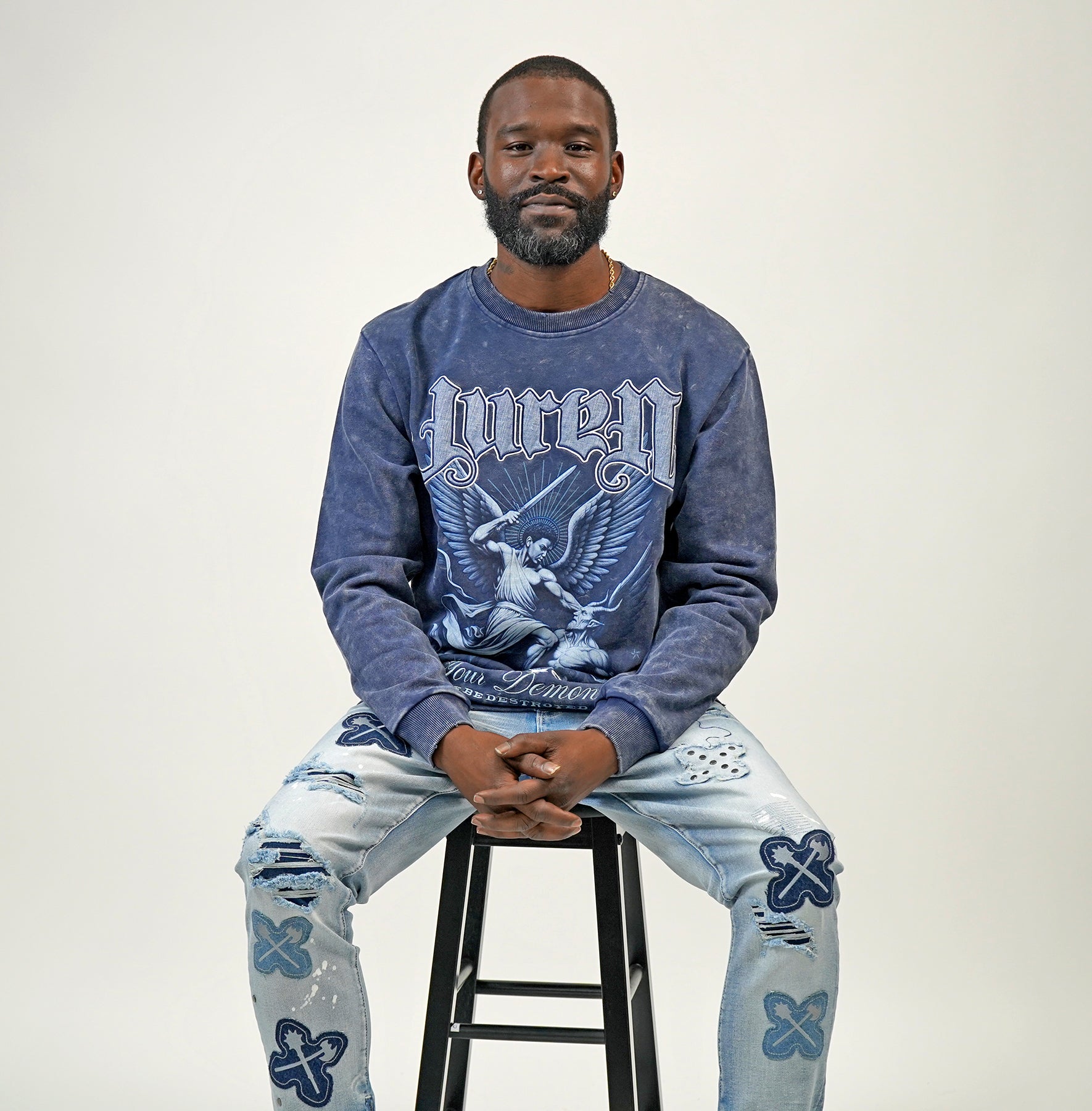 Man wearing a blue graphic sweatshirt and light blue jeans sitting on a black stool against a white background
