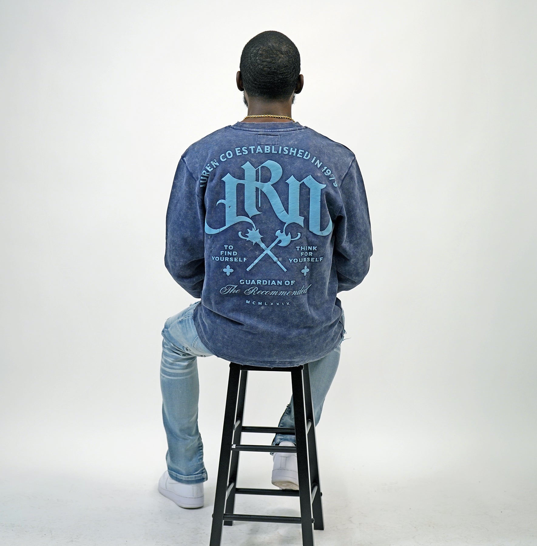 Person wearing a blue acid wash sweatshirt with text on the back, sitting on a black stool against a white background