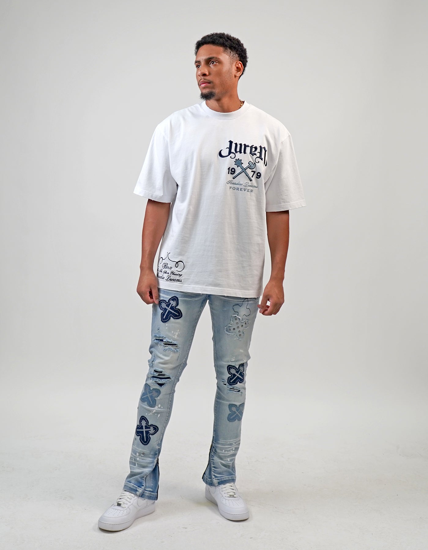 Person wearing a white t-shirt with text and graphics, paired with light blue jeans featuring floral patterns on a plain background. White t-shirt with navy text and graphics on a white shirt "Juren 1979 Logo.
