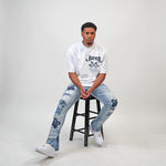 Person wearing a white t-shirt and blue jeans sitting on a black stool against a gray background. Person wearing a white t-shirt with text and graphics, paired with light blue jeans featuring floral patterns on a plain background. White t-shirt with navy text and graphics on a white shirt "Juren 1979 Logo.