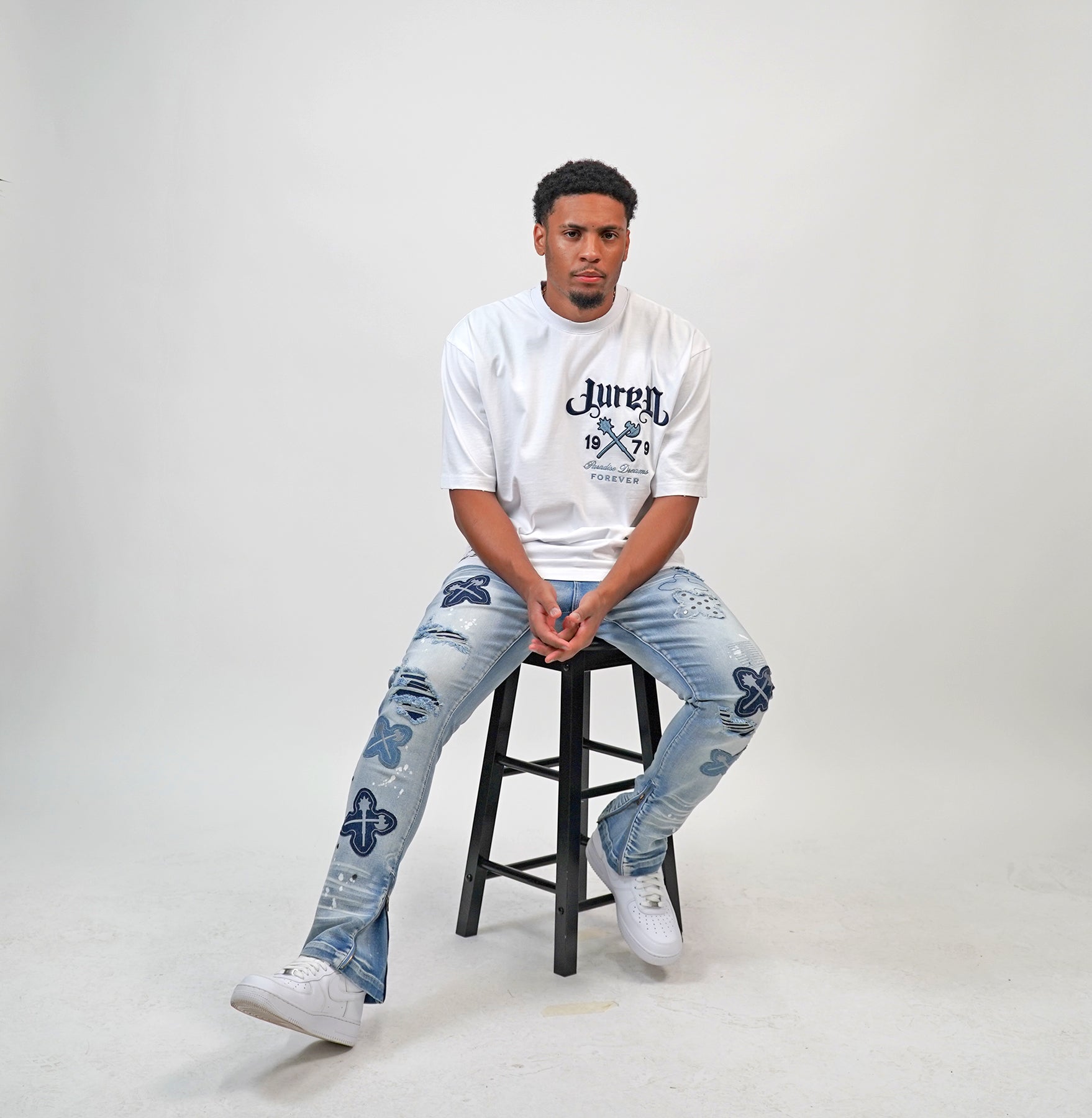 Person wearing a white t-shirt and blue jeans sitting on a black stool against a gray background. Person wearing a white t-shirt with text and graphics, paired with light blue jeans featuring floral patterns on a plain background. White t-shirt with navy text and graphics on a white shirt "Juren 1979 Logo.