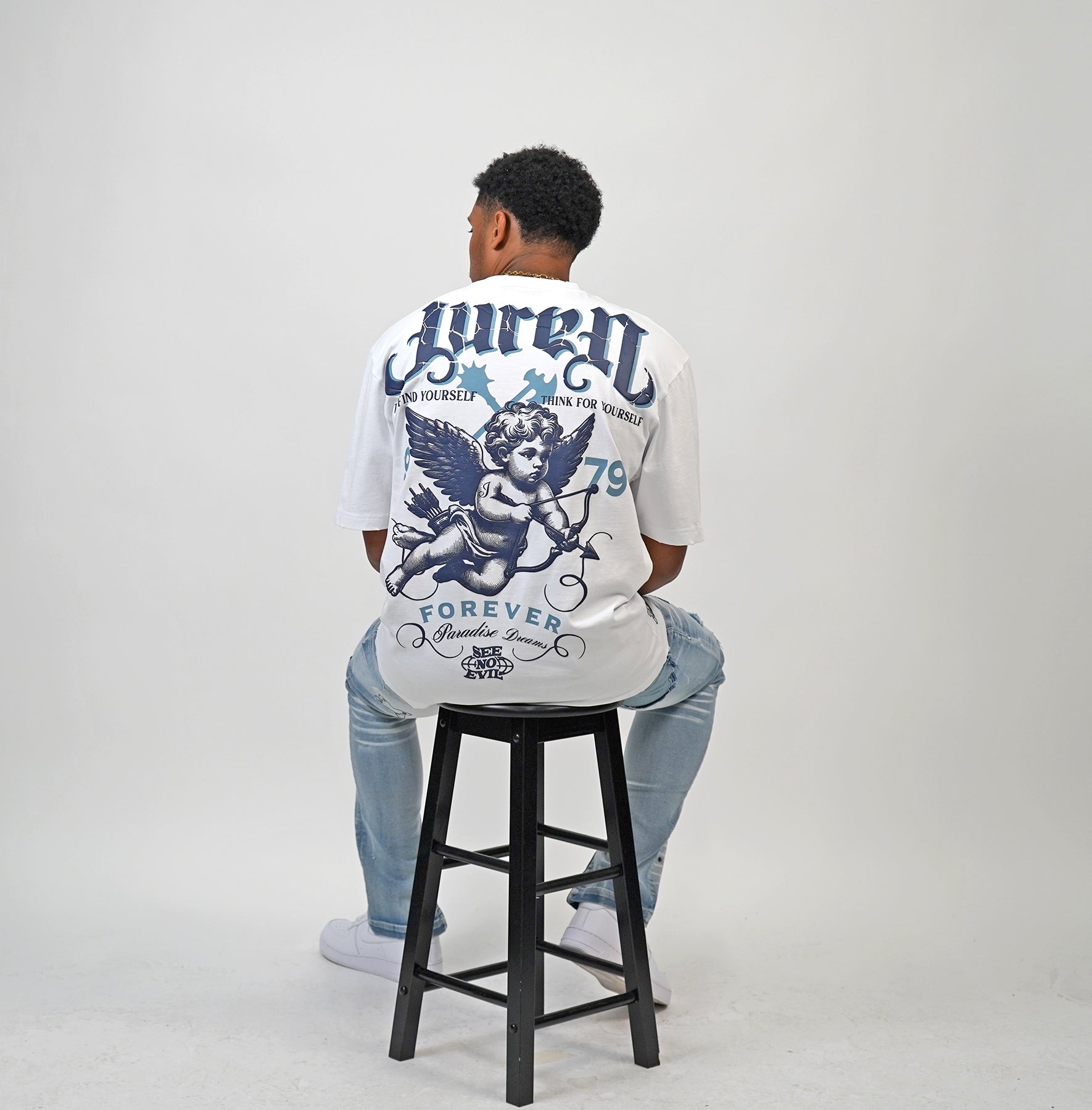 Person wearing a white t-shirt with Juren Logo on top with an Angel graphics and text, sitting on a black stool against a plain background